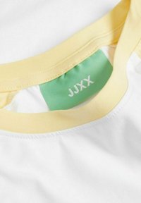 Camiseta con un cuerpo blanco y un cuello amarillo. Presenta una etiqueta verde con "JJXX" en blanco. Textura de tela suave y lisa.