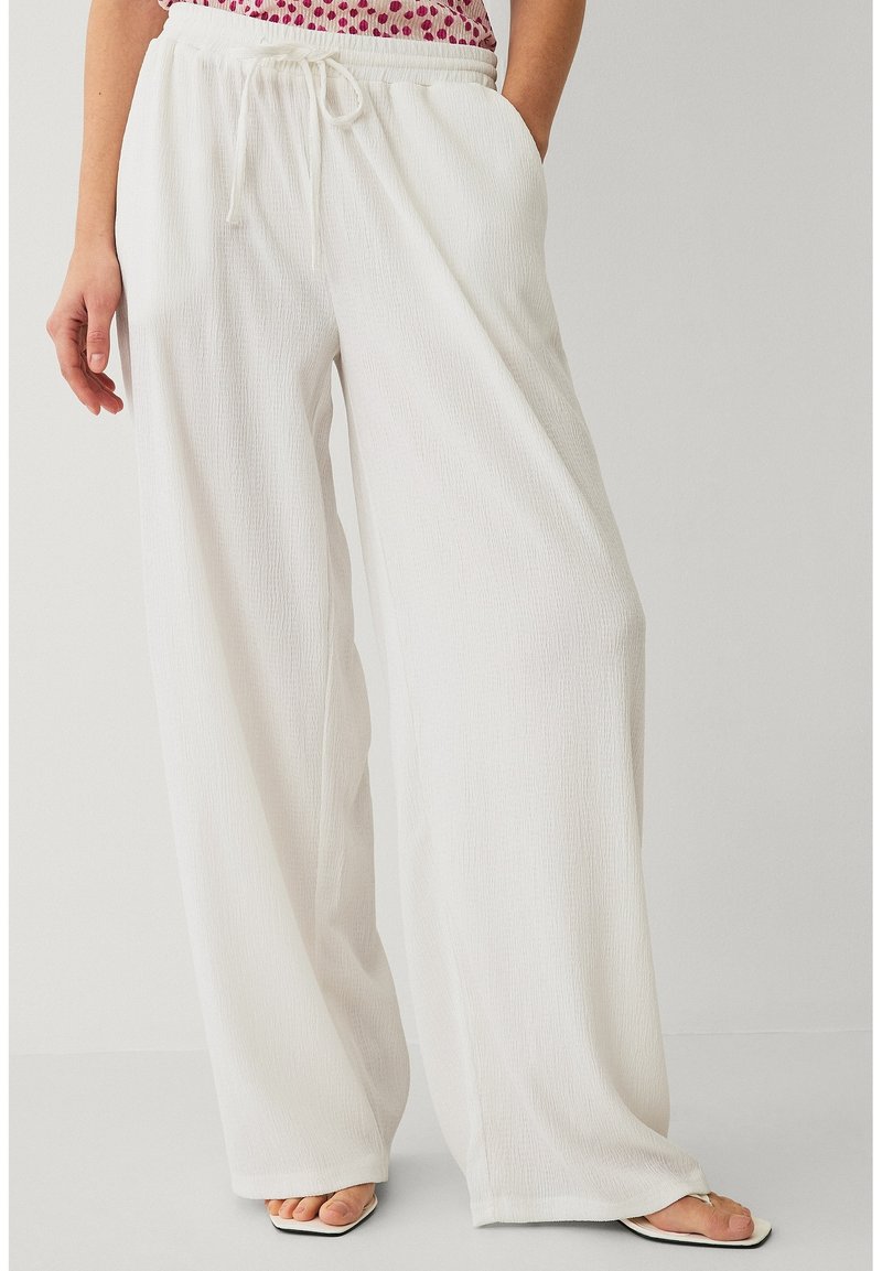 Vrouw draagt losse witte broek met trekkoord, brede pijpen en vierkante sandalen, staand tegen een effen achtergrond.