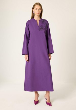 Femme portant une longue robe violette avec une broderie florale sur l'épaule et des talons hauts violets assortis, debout sur un fond uni.