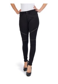 Mujer con pantalones skinny negros con cremallera y detalles de rodilla acanalados, combinados con tacones altos negros y una blusa blanca.