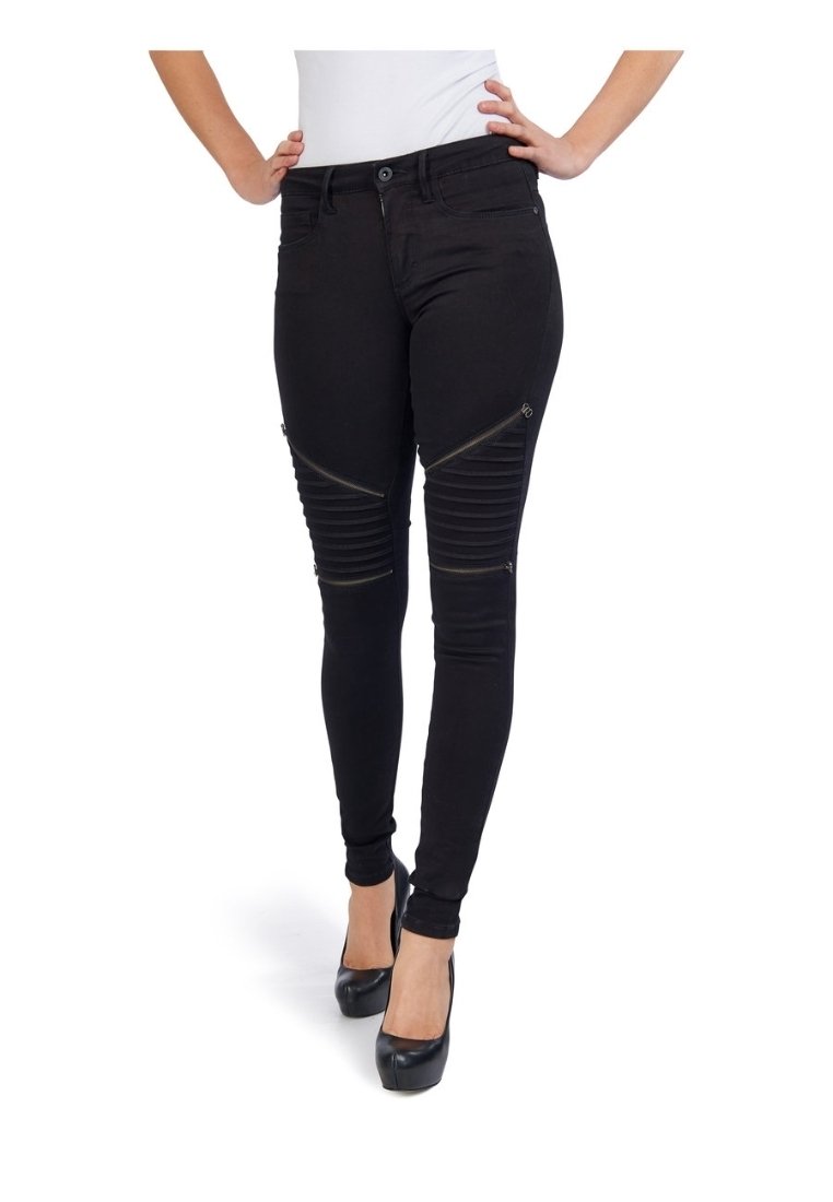 Mujer con pantalones skinny negros con cremallera y detalles de rodilla acanalados, combinados con tacones altos negros y una blusa blanca.