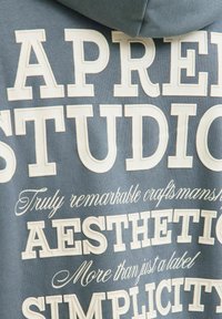 Šedá mikina s bílým embosovaným textem, včetně "APPAREL" a "STUDIO." Měkká textura, pohodlný design a s kapucí.