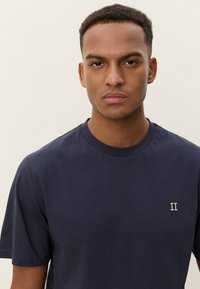 Tamsiai mėlynas medvilninis marškinėlis trumpomis rankovėmis, su apvalia iškirpte ir mažu baltu siuvinėtu "II" simboliu kairėje krūtinės pusėje. Lygus paviršius.