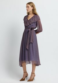 Paarse en rode jurk met een patroon, een ruches halslijn, lange mouwen, een strikceintuur en een vloeiende zoom. Bruine hakken maken de look compleet.