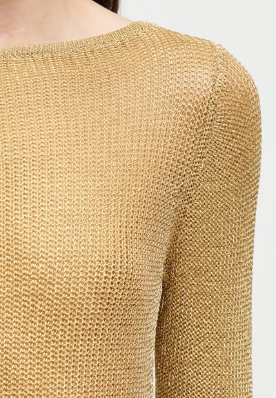 Κοντινό πλάνο ενός ατόμου που φοράει χρυσαφένιο, λεπτοδουλεμένο μεταλλικό μπλουζάκι με μακριά μανίκια και στρογγυλή λαιμόκοψη.