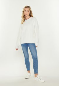Femeie îmbrăcată într-un pulover alb din tricot, blugi skinny albaștri și adidași albi, în fața unui fundal simplu și deschis.