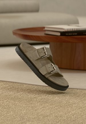 Sandalo in camoscio beige con due fibbie metalliche regolabili e suola nera, posizionato su un tappeto beige strutturato vicino a un tavolino da caffè in legno e libri.
