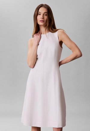 Kobieta z długimi brązowymi włosami, ubrana w bezrękawową, jasnobeżową sukienkę do kolan, stojąca na tle jednolitego szarego tła.