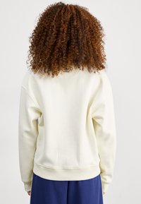 Vit sweatshirt med strukturerad textil, vida ärmar och ribbad nederkant. Har en liten logotyp på baksidan av halsringningen.