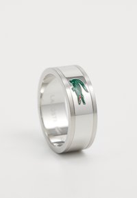 Sidabrinės spalvos metalinis žiedas su lygiu paviršiumi, centre kuris yra žalios ir raudonos spalvos krokodilo logotipas. Paprastas ir modernus dizainas.