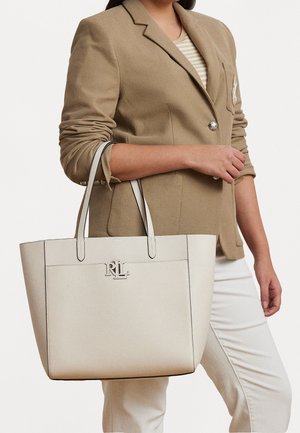 Femme portant un blazer beige et un pantalon blanc, tenant un grand sac fourre-tout en cuir blanc cassé avec le logo RL sur le devant.