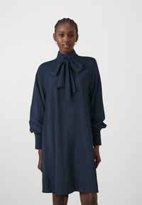 Laivansinis mekko, jossa on korkea kaula, nauhakiinnitys ja pitkät puffihihat. Valmistettu sileästä kankaasta, jonka siluetti on löysä ja suora.