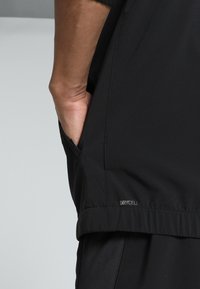Top sportiv negru cu o textură netedă, având un buzunar pe lateral și branding "DRYCELL" pe tiv. Mânici scurte și un design lejer.
