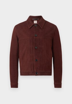 Férfi barna, gombos dzsekiben, szürke galléros pólóban, bézs nadrágban fekete övvel és fehér tornacipőben, egyszínű háttér előtt áll.