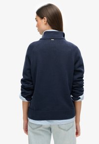 Mujer con cabello largo y castaño, vestida con un suéter de punto azul marino con las mangas remangadas sobre una camisa azul clara y jeans de lavado claro, vista de espaldas.