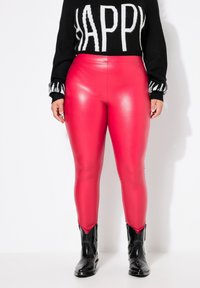Leggings aderenti in pelle sintetica rossa con finitura lucida, abbinati a un maglione nero con scritte bianche in grassetto e stivali neri alla caviglia.