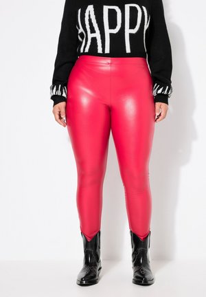 Figurbetonte rote Kunstlederleggings mit glänzender Oberfläche, kombiniert mit einem schwarzen Pullover mit fettgedrucktem weißen Schriftzug und schwarzen Ankle Boots.