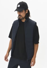 Navy puffer vest with a high collar, zip closure, and quilted texture worn over a black t-shirt; model wearing a black cap with logo.