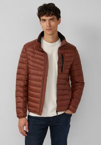 Kastanjebruine pufferjas met een ritssluiting aan de voorkant, voorzien van een staande kraag, quilted textuur en zijzakken. Draag het over een crèmekleurige trui en jeans.