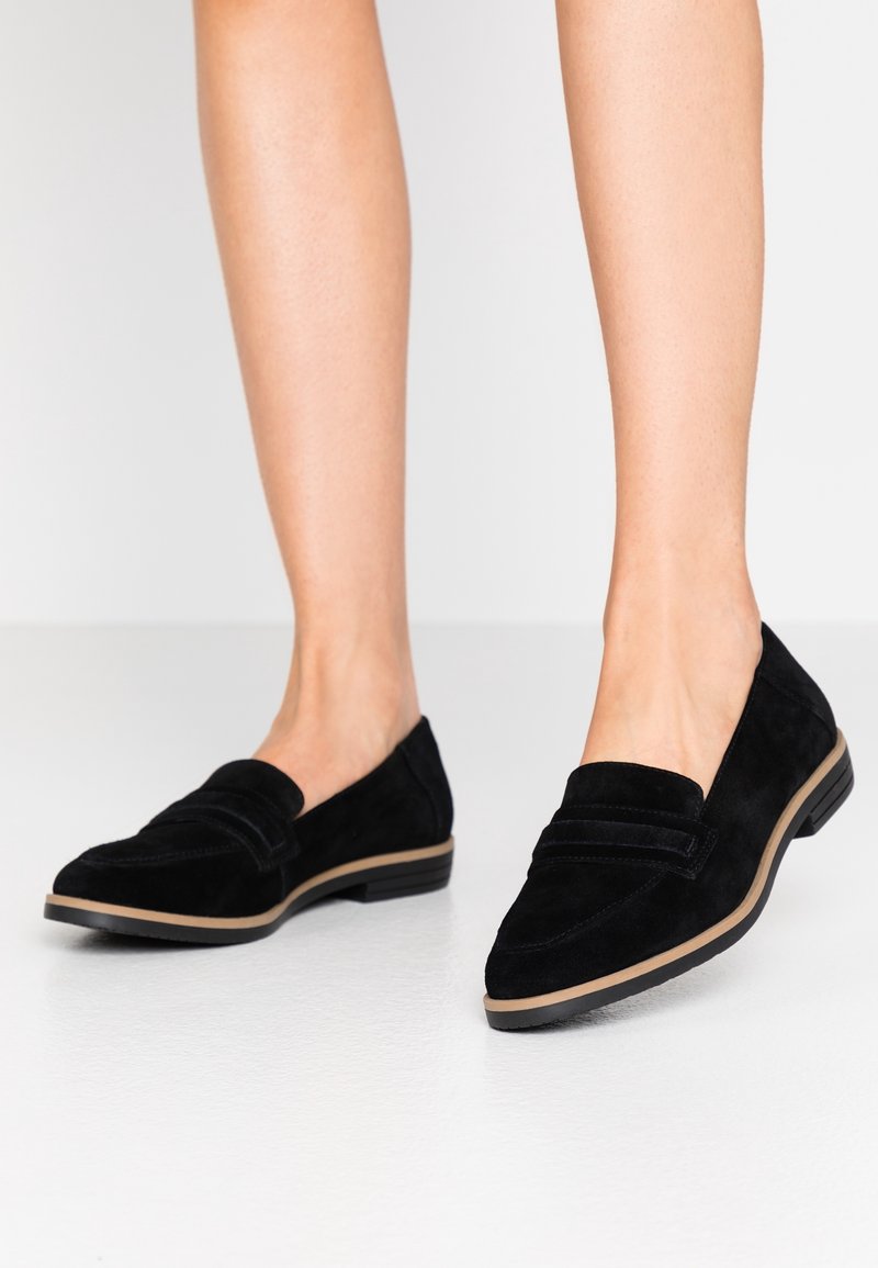 Mocassins en daim noir avec un petit talon, un bout pointu, et des rayures décoratives sur la tige. La semelle présente un bord contrastant de couleur marron clair.