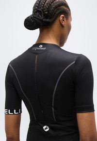 Άτομο με πλεξούδες σε χαμηλό κότσο, φοράει μαύρη φανέλα ποδηλασίας με ανακλαστικές ραφές και λευκά λογότυπα στην πλάτη και στο μανίκι.