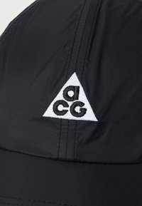 Black cap featuring a triangular white patch with embroidered "acg" logo. Made of lightweight material with visible stitching details.