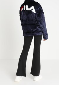 Marinblå fuskpälsjacka med en stor vit och röd Fila-logotyp på ryggen. Matcher med svarta utsvängda byxor och vita sneakers.