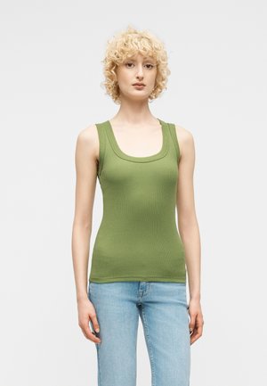 Γυναίκα με σγουρά ξανθά μαλλιά, ντυμένη με στενό αμάνικο τοπ ελιάς και ανοιχτόχρωμα μπλε τζιν, στέκεται μπροστά σε απλό ανοιχτόχρωμο φόντο.