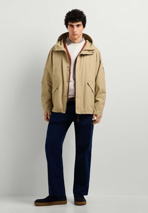 Jonge man in een beige jas met capuchon, witte trui, geruit overhemd, donkerblauwe broek en zwarte schoenen, staand tegen een effen lichte achtergrond.