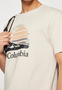 Bežové tričko s krátkým rukávem, na kterém je barevná grafika západu slunce s horizontálními pruhy a siluetami hor pod slovem "Columbia."