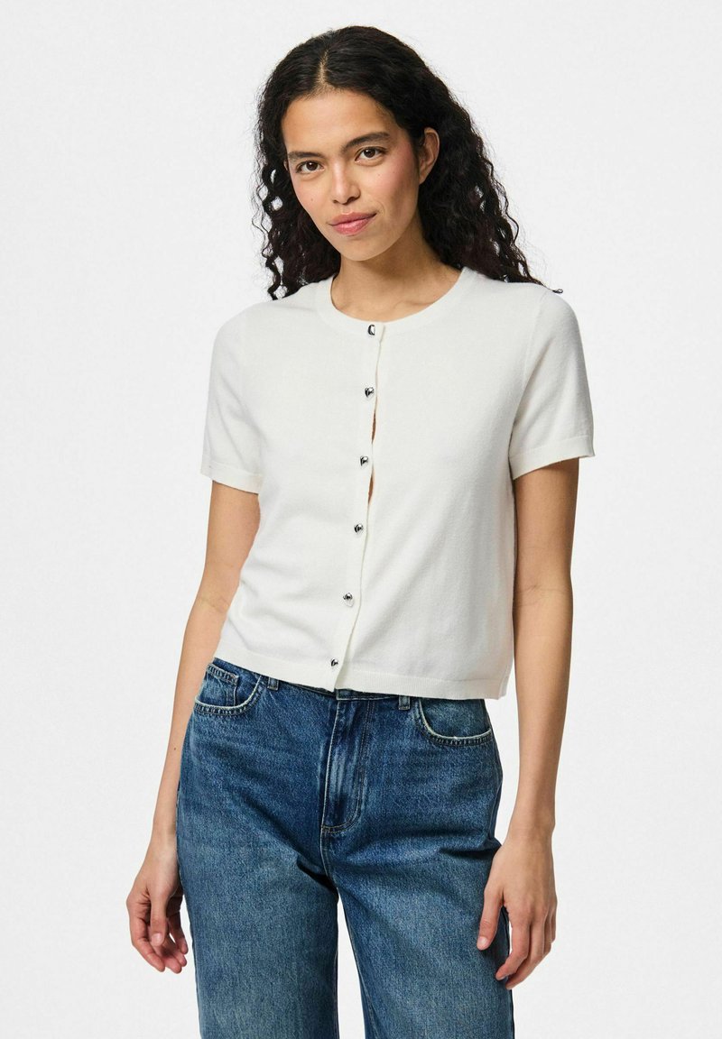 Lätt kortärmad vit cardigan med rund halsringning och silverknappar framtill, tillverkad av mjukt tyg och tillsammans med blå jeans.