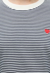 Camiseta de rayas horizontales blancas y negras con un pequeño corazón rojo bordado en la parte derecha del pecho, debajo del cuello.