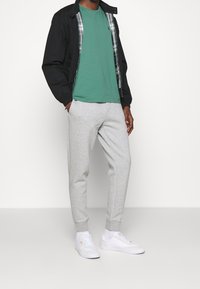 Man som bär svart jacka, grön t-shirt, ljusgråa sweatpants, vita sneakers, står med händerna i fickorna mot en vit bakgrund.