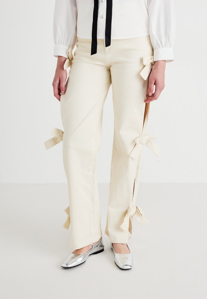 Pantalones de color crema con aberturas laterales, adornados con lazos de tela. La tela parece ligera y suave. Combinados con zapatos planos plateados.