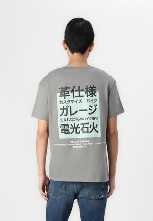 Man draagt een grijs T-shirt met Japanse tekst en Engelse zinnen over Replay Garage op de rug, gecombineerd met een blauwe spijkerbroek.