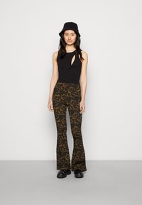 Top preto sem mangas com detalhe recortado, combinado com calças castanhas de boca de sino com padrão de leopardo escuro. Chapéu de balde preto e calçado.
