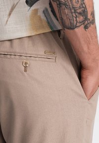 Pantaloni beige chiaro, in misto cotone, con una tasca posteriore con bottone e un piccolo tag metallico del brand. Tessuto liscio, vestibilità tailored.