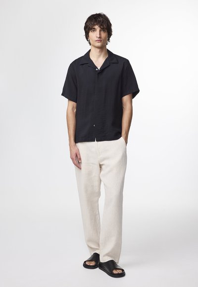 Svart kortärmad skjorta i strukturerad tyg, kombinerad med ljus beige linnebyxor och svarta slip-on sandaler. Enkel design med öppen krage.