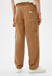 Bawełniane spodnie cargo w jasnobrązowym kolorze, z wieloma kieszeniami, przeszyciami i luźnym krojem, zestawione z białymi sneakersami.