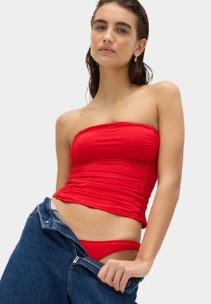 Kobieta ubrana w czerwony top bez ramiączek i pasujące czerwone spodnie, ściąga niebieskie dżinsy, patrząc w kamerę z neutralnym wyrazem twarzy.