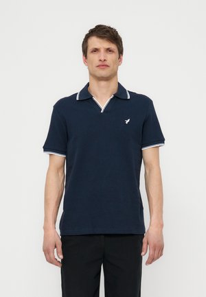 Άνδρας που φοράει μπλε σκούρο πόλο με λευκά περιθώρια στον γιακά και τα μανίκια, στέκεται μπροστά σε απλό φωτεινό φόντο.