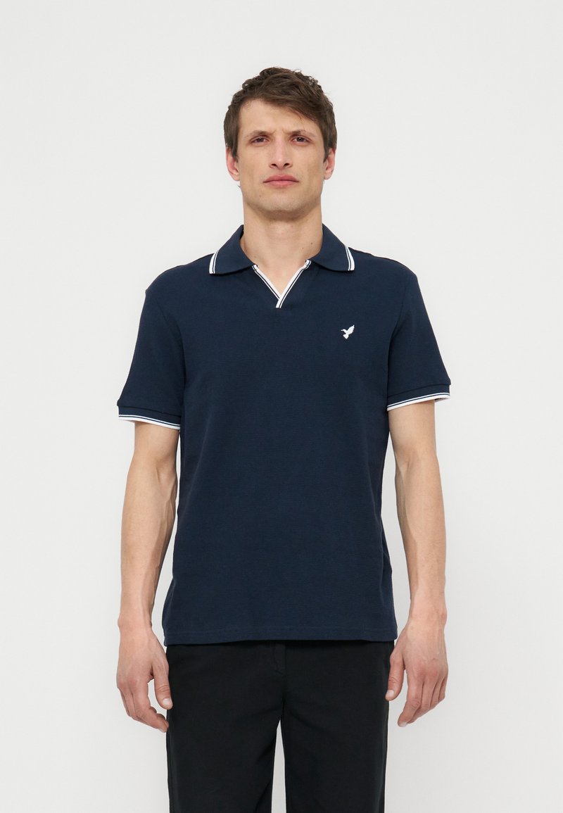 Άνδρας που φοράει μπλε σκούρο πόλο με λευκά περιθώρια στον γιακά και τα μανίκια, στέκεται μπροστά σε απλό φωτεινό φόντο.