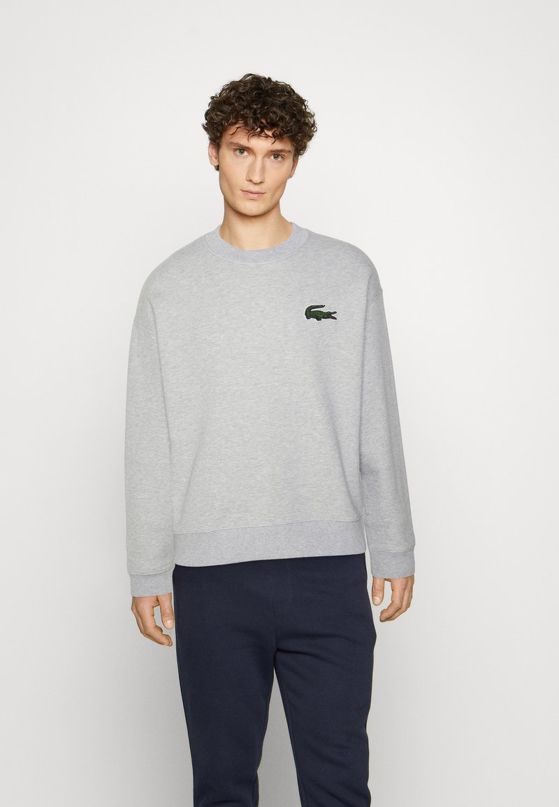 Szary sweatshirt z prążkowanymi mankietami i dołem, z małym, zielonym, haftowanym logo krokodyla po lewej stronie klatki piersiowej. Gładka tekstura materiału.