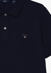 Marinblå piké skjorta i texturerat tyg, med en treknapps placket och en liten broderad GANT-logotyp på bröstet.