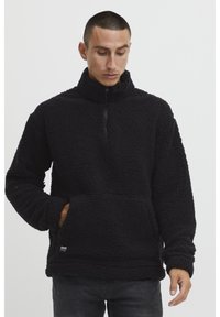 Pullover in pile nero con colletto alto, chiusura a zip quarto di lunghezza e tasca frontale. Esterno morbido e strutturato con una vestibilità rilassata.
