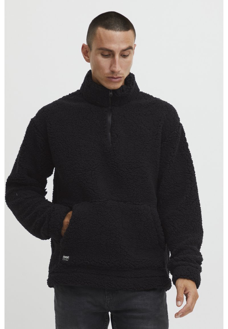Pullover in pile nero con colletto alto, chiusura a zip quarto di lunghezza e tasca frontale. Esterno morbido e strutturato con una vestibilità rilassata.