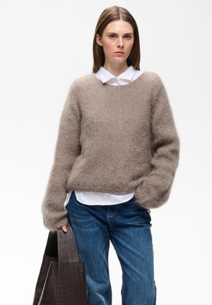 Ung kvinde iført en beige fnugget sweater, hvid skjorte og blå jeans, der holder en stor mørkebrun tekstureret tote-taske.