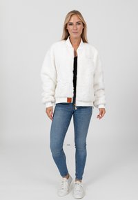 Weiße Kunstfell-Bomberjacke mit ribbelten Bündchen und Saum, Frontreißverschluss und einem orangefarbenen Label-Patch, kombiniert mit blauen Jeans und weißen Sneakers.
