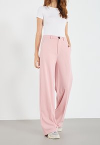 Woman wearing high-waist wide-leg pink trousers, white fitted short-sleeve top, and white sneakers, standing with one hand in pocket.