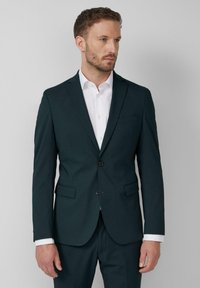 Blazer ajustado de color verde oscuro con solapas de muesca, cierre de un solo botón, dos bolsillos frontales y una textura suave. Combinado con una camisa blanca.